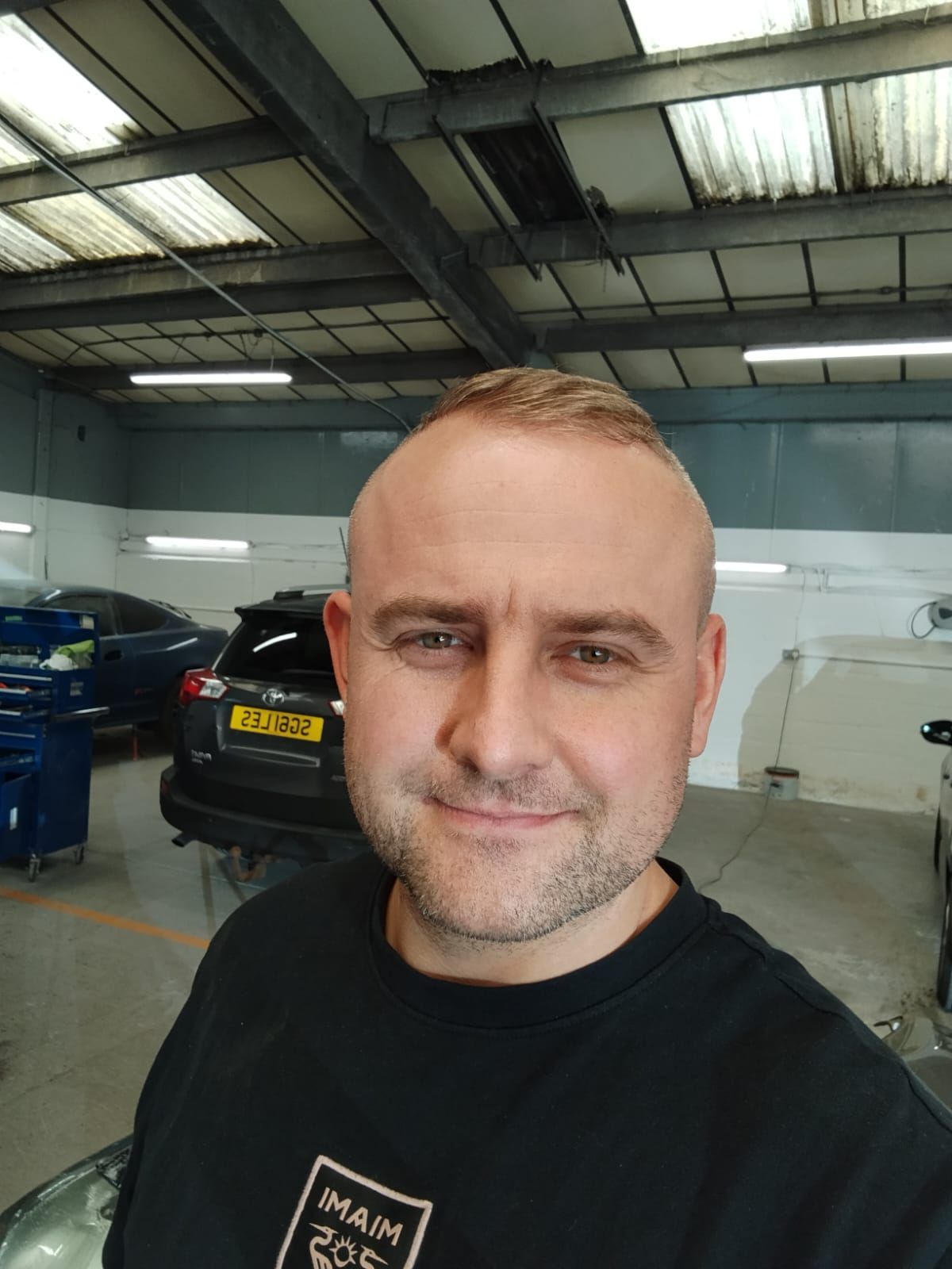 Man taking a selfie in an auto garage with cars and work lights in the background
