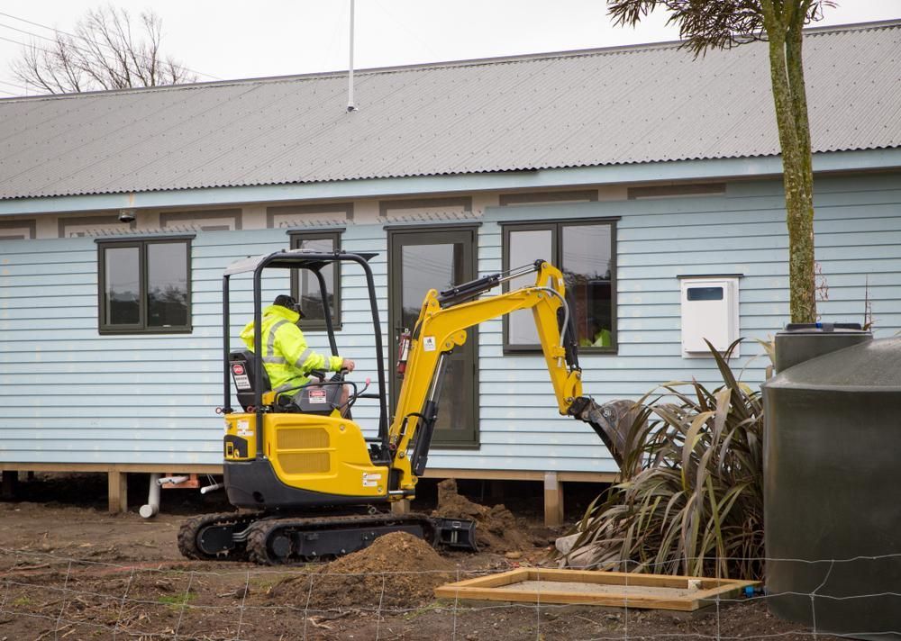 A Man is Driving a Small Yellow Excavator in Front of a House — Husky Demolition In Waratah, NSW