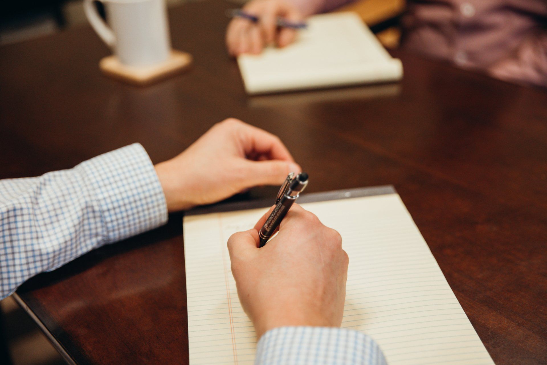 Hands writing in a notebook at a meeting table with another notebook and a blurred person in the background