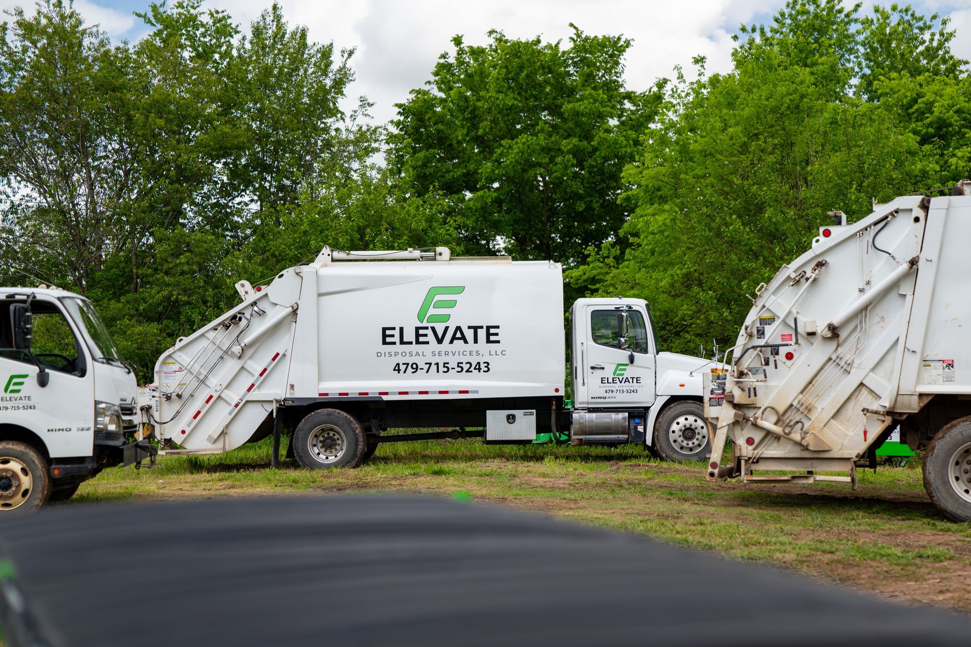 White Elevate garbage truck parked beside other waste trucks on a grassy roadside, trees in background