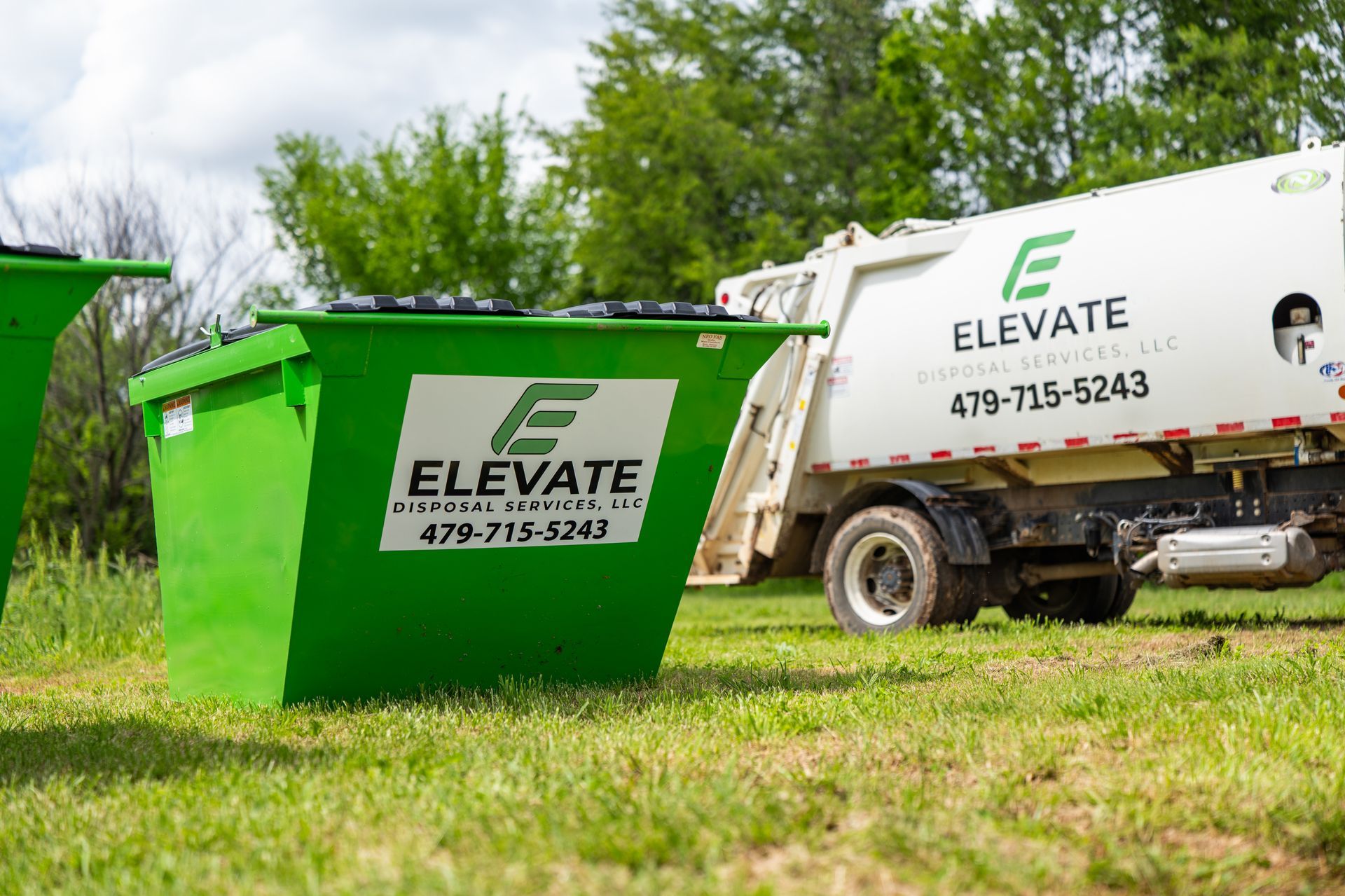 Green ELEVATE dumpster and white waste truck parked on grass in a wooded area.