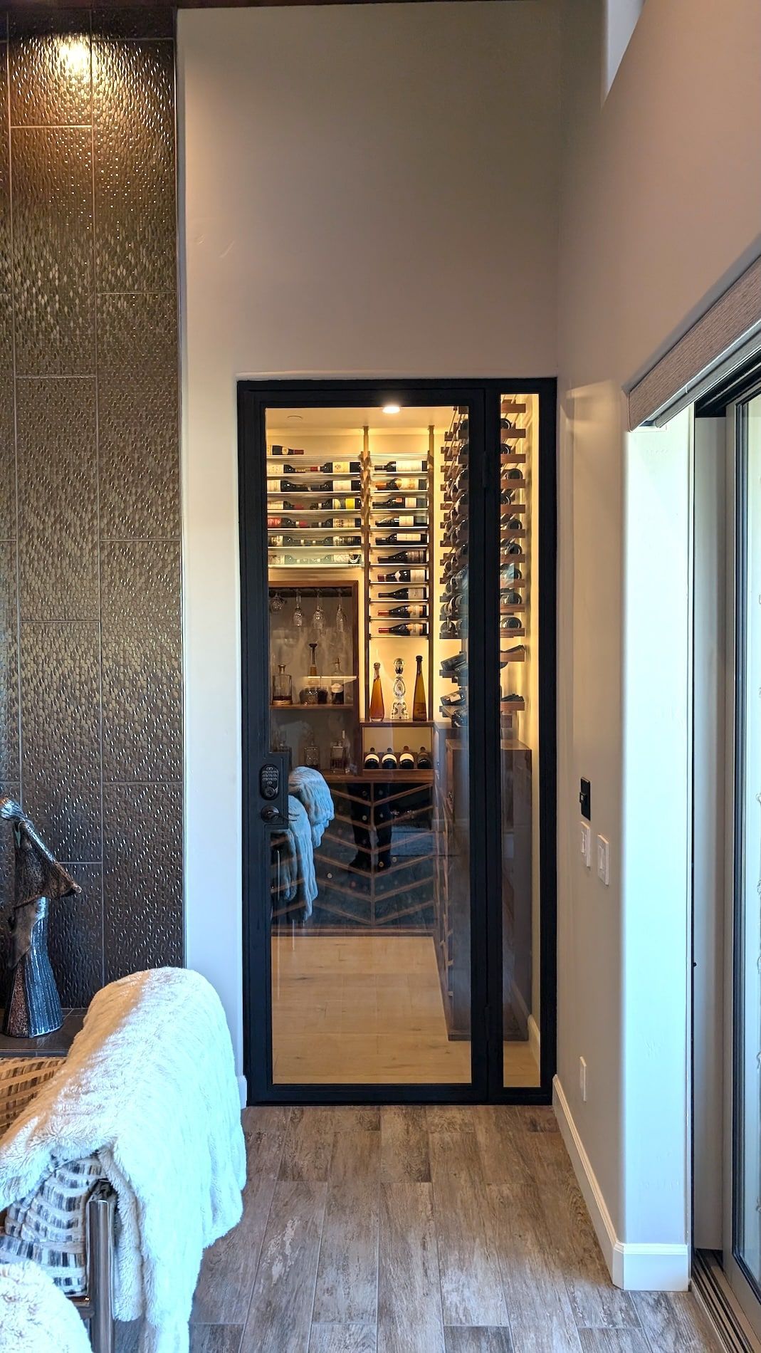 Modern bathroom with glass shower door, stone tile walls, and window light showing a lit wine room beyond