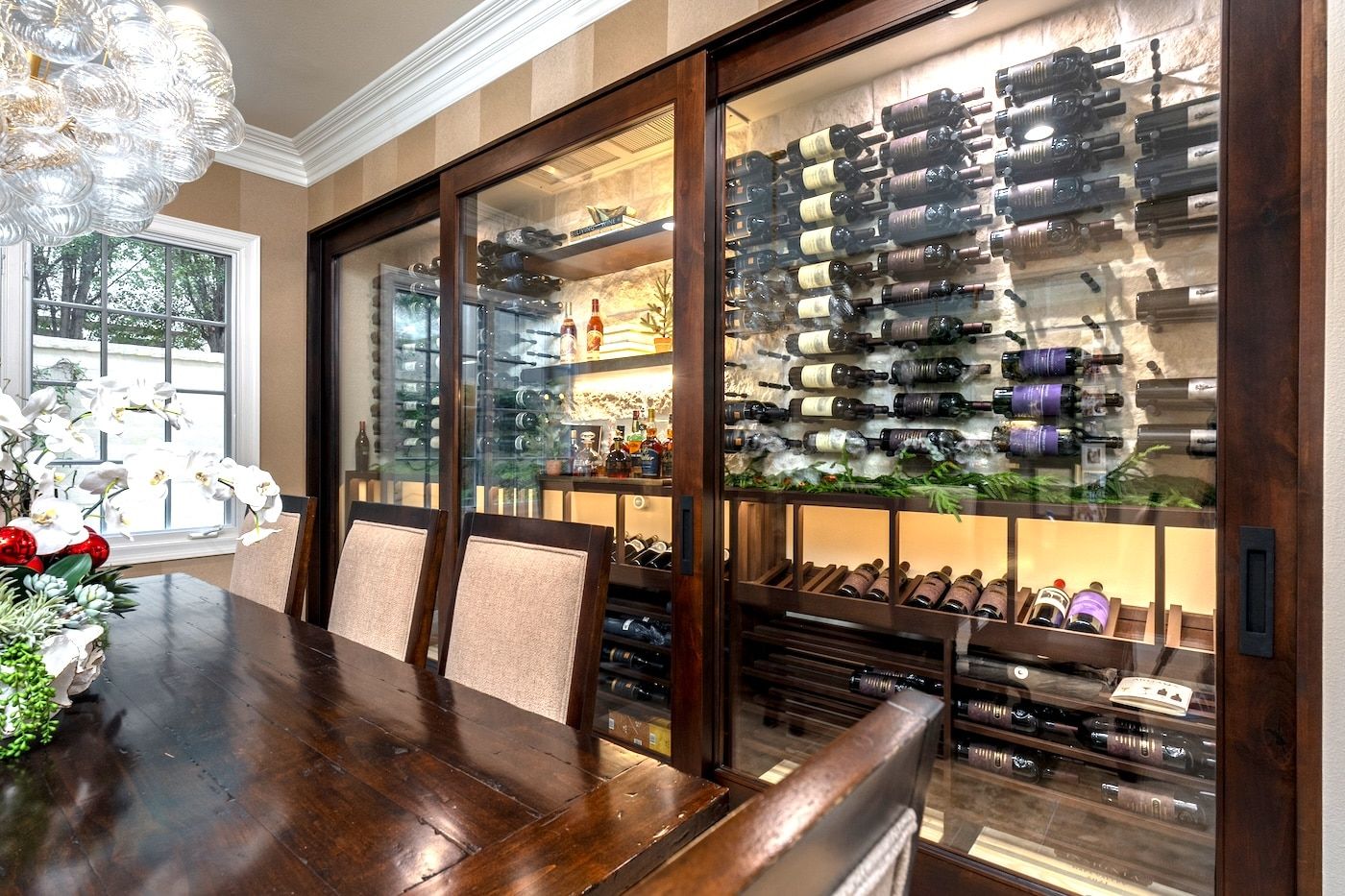 Dining room with a glass-front wine cabinet, bottles stacked on shelves, and a wooden table by a window.