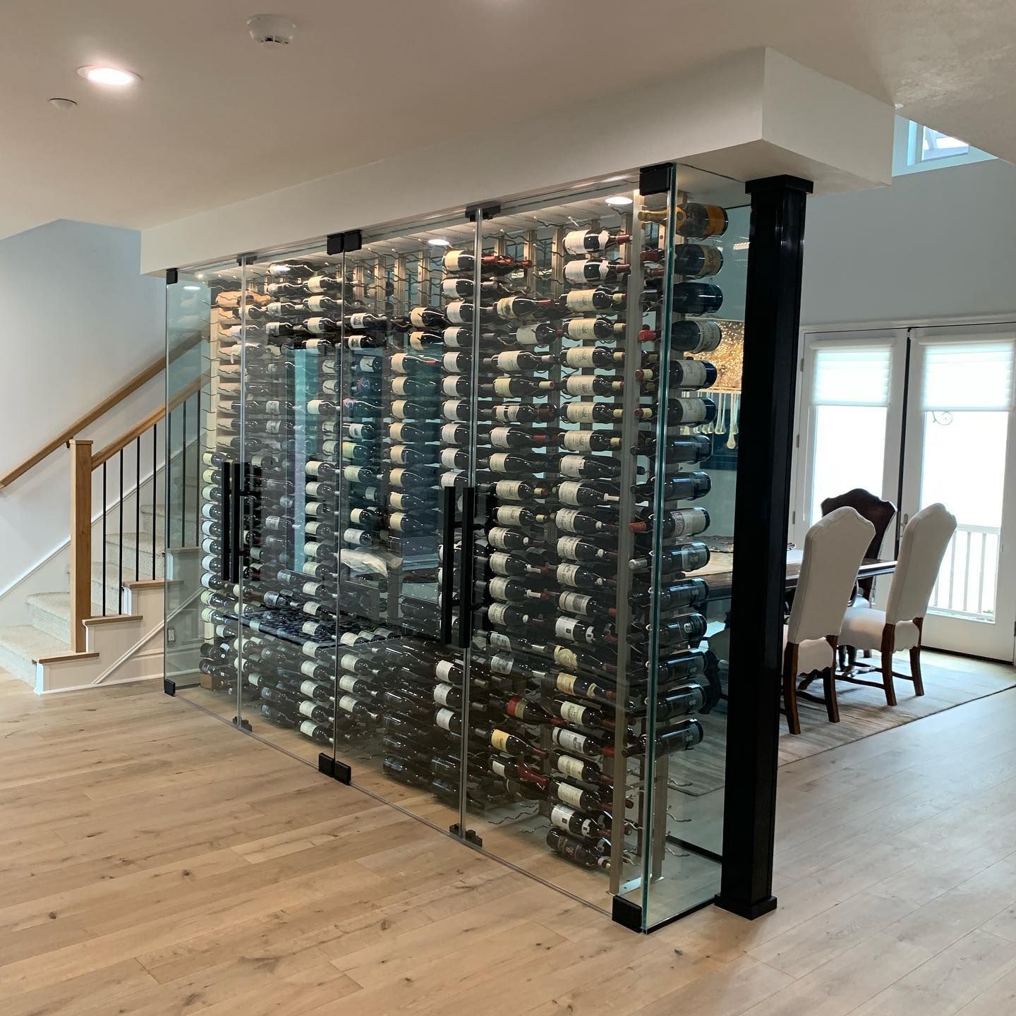 Glass wine cellar divider in a modern home, filled with wine bottles beside a staircase and dining area.