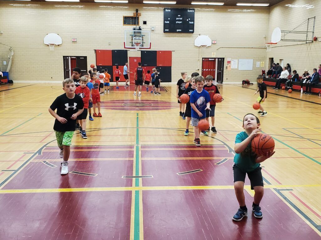 Children playing basketball in a gym, with one boy dribbling in the foreground and others running behind.