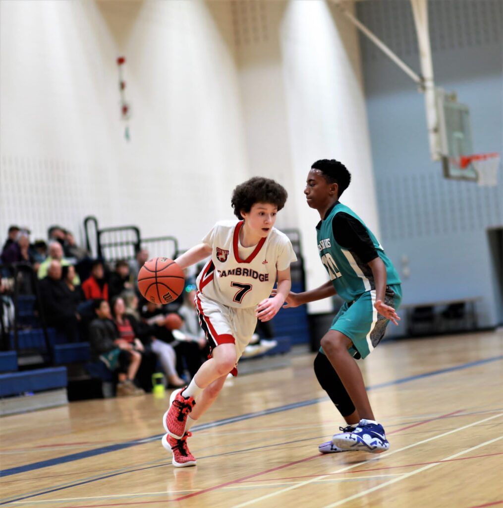 Two basketball players dribble and defend on an indoor court, with spectators in the background.