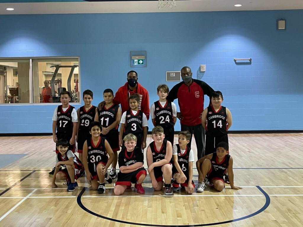 Youth basketball team posing on a gym court, wearing red and black uniforms, with coaches in back