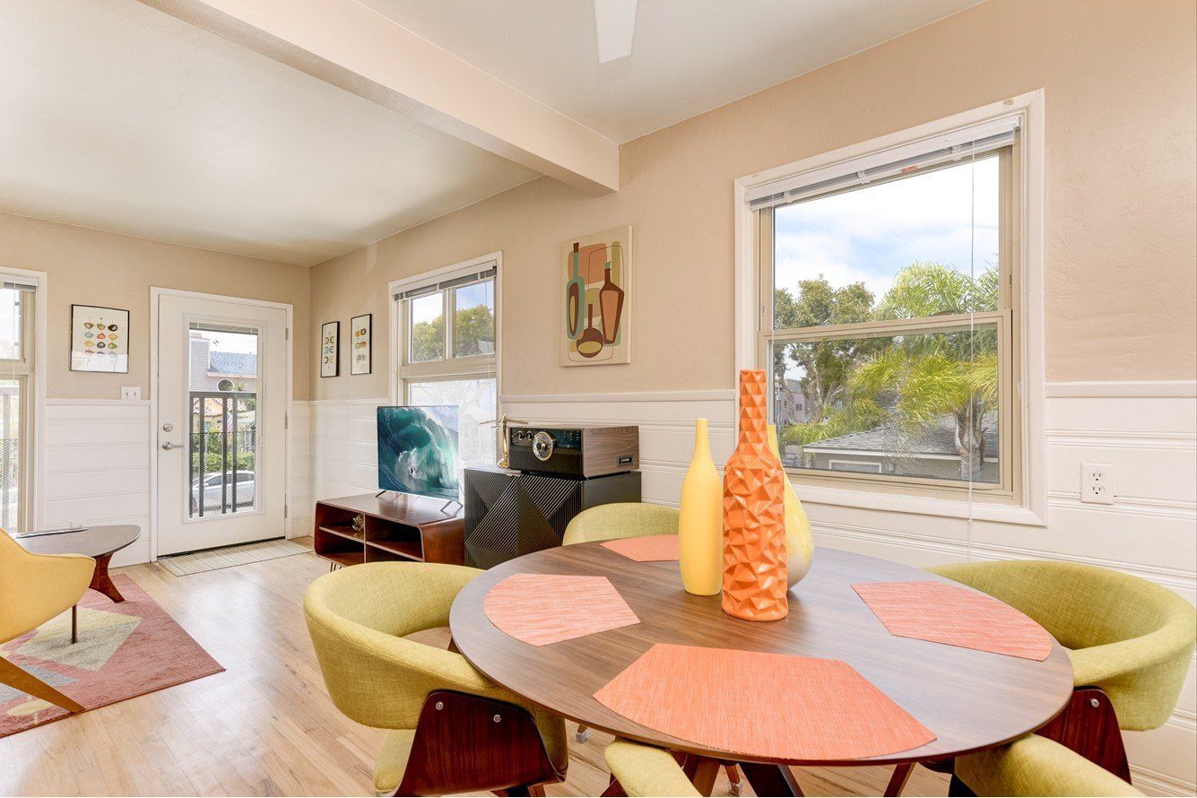 A bright dining room with round table and mid-century chairs, tall orange vase, and a view through the window.