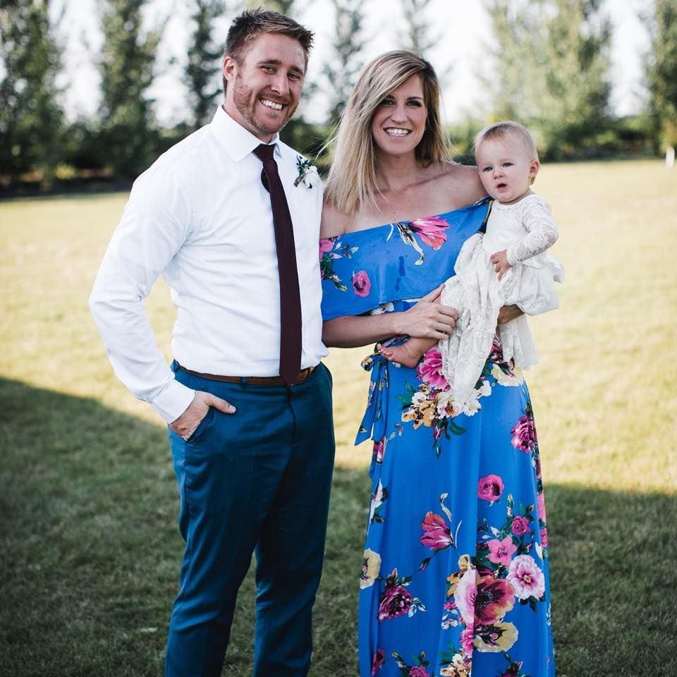 A man in a white shirt and tie is standing next to a woman in a blue floral dress holding a baby.