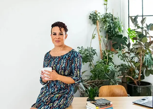Person in a patterned dress holding a mug beside a laptop and plants in a bright room