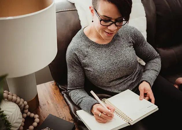 Person writing in a notebook while seated on a couch beside a lamp