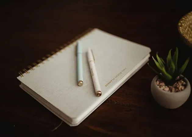 Minimal desk scene with a white notebook, two pencils, and a small potted plant on a dark wooden table