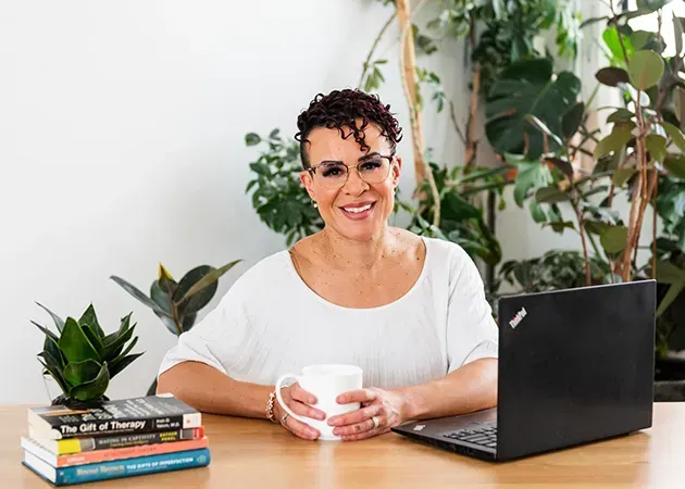 Woman at a desk holding a mug beside a laptop and stacked books, with plants in the background.