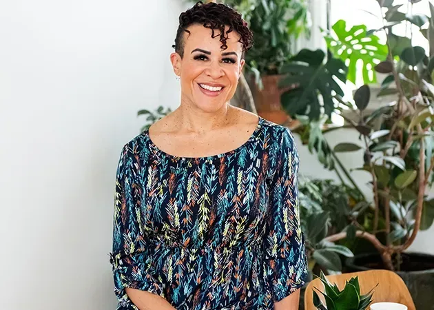 Smiling person in a colorful blouse seated indoors with a large leafy plant nearby