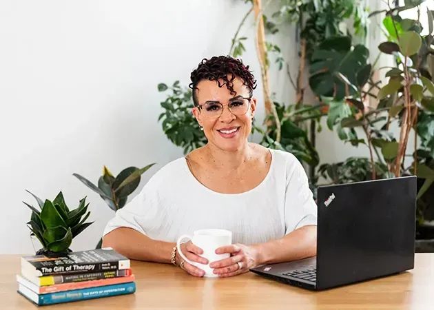 Person smiling at a desk with a laptop, mug, potted plants, and stacked books in a bright room