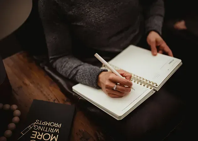 Person writing in a spiral notebook on a couch, with a book beside them.