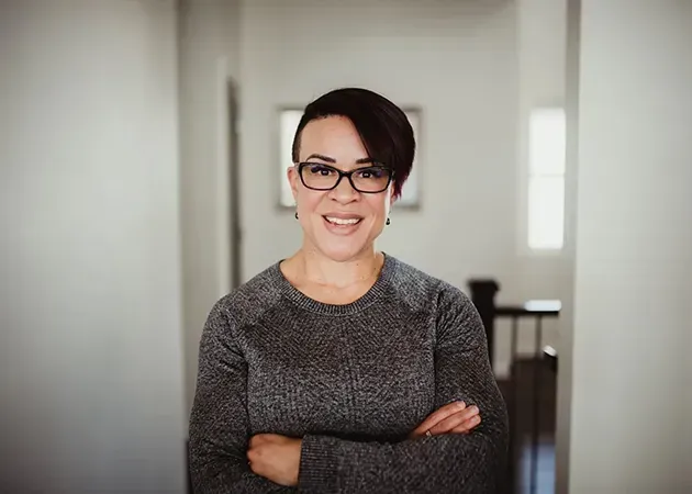 Smiling person with glasses and crossed arms in a gray sweater, standing in a bright hallway