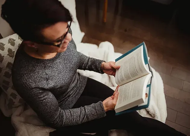 Person reading an open book while seated on a couch in a cozy indoor setting