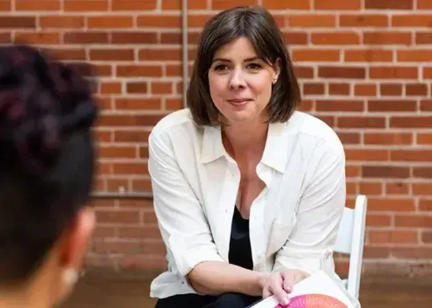Woman in a white shirt sitting and speaking against a brick wall backdrop