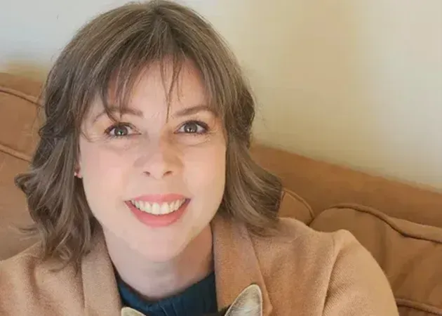 Smiling woman with short brown hair sitting on a tan couch, wearing a tan coat and blue top.