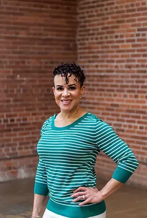 Smiling person in a teal striped shirt, standing with hand on hip against a brick wall.