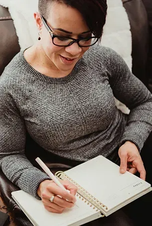 Woman in glasses writing in a notebook while sitting on a couch