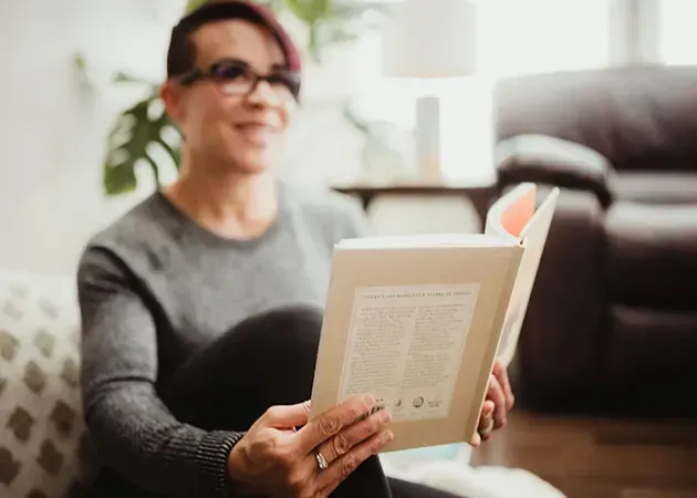 Person with glasses reading a book on a couch in a bright living room