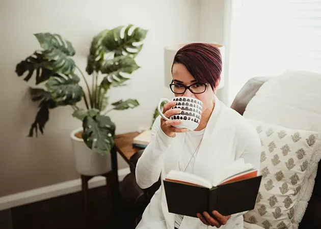 Person reading a book while sipping tea in a bright living room beside a potted plant