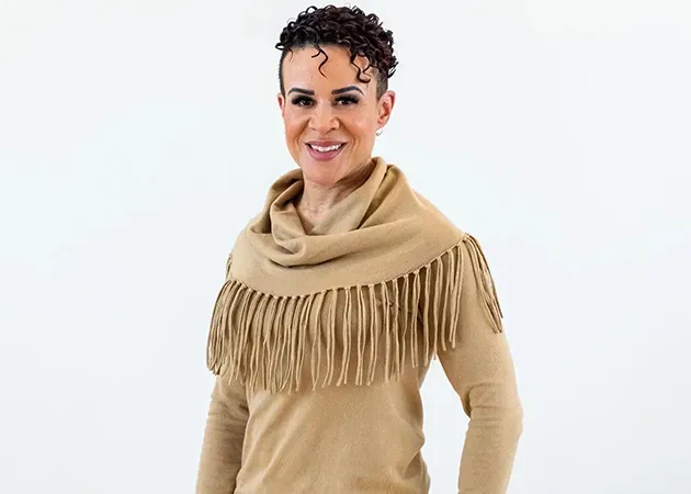 Woman in a beige fringed shawl sweater smiling against a white background