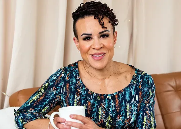 Woman in a teal patterned blouse smiling while holding a white mug on a tan couch
