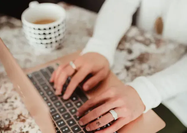 Hands typing on a laptop beside a coffee cup on a light-colored desk
