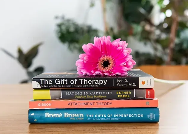 Stack of therapy and psychology books with a pink flower on top, on a table with a blurred plant background