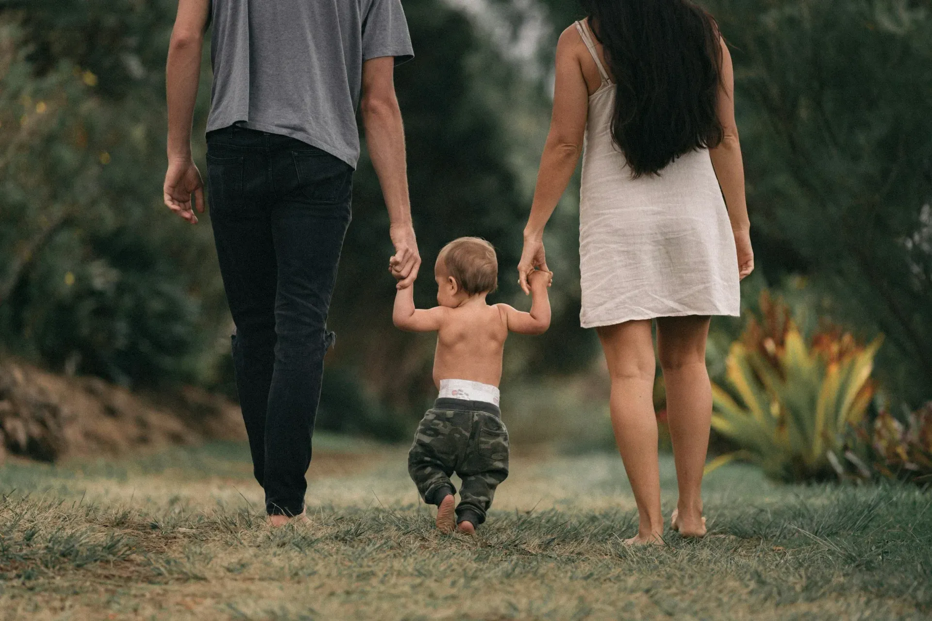 Three people walking away on a grassy path, holding hands, with a toddler between two adults.