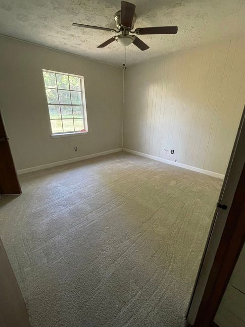 Empty carpeted bedroom with off-white walls, a window, and a ceiling fan.