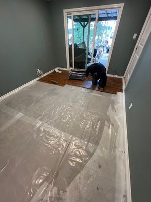 Worker kneeling on hardwood floor beside sliding glass door, preparing a room with protective plastic sheeting.