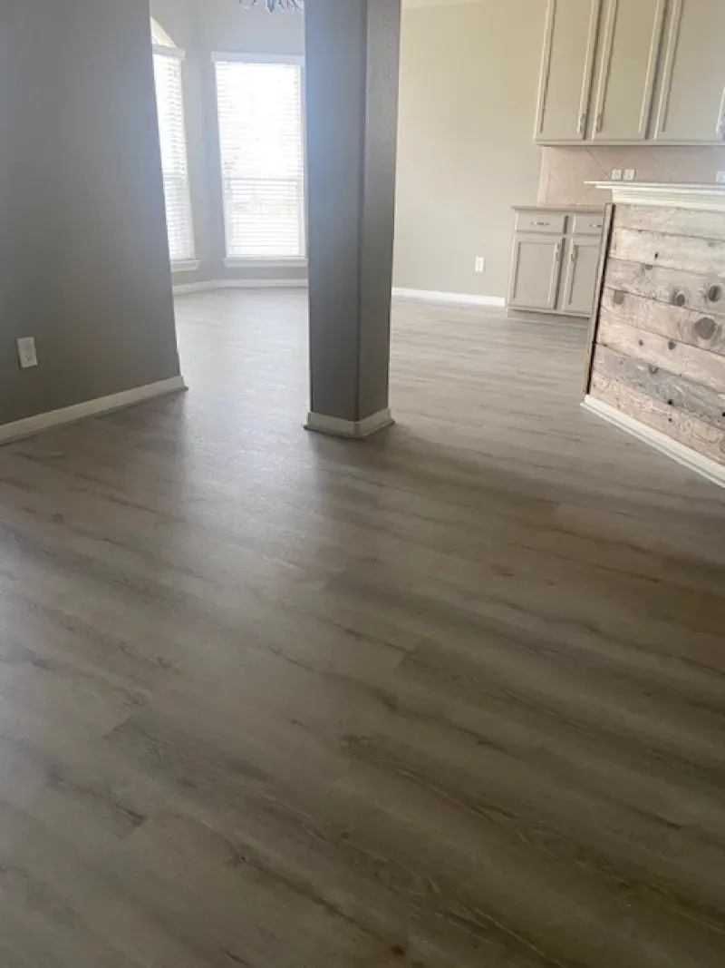 Empty room with light wood flooring, gray walls, central support column, and a fireplace by bright windows.