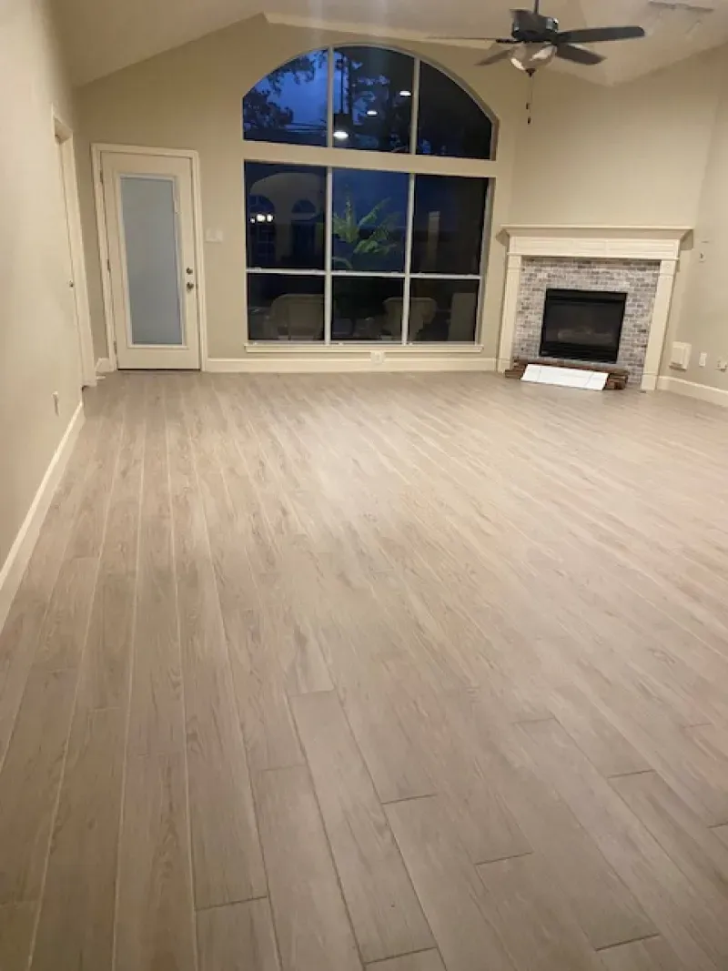 Empty living room with light wood flooring, large arched window, ceiling fan, and stone fireplace.