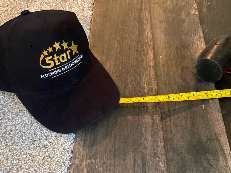 Black cap with gold logo beside a yellow measuring tape on a wooden floor with a chair leg nearby