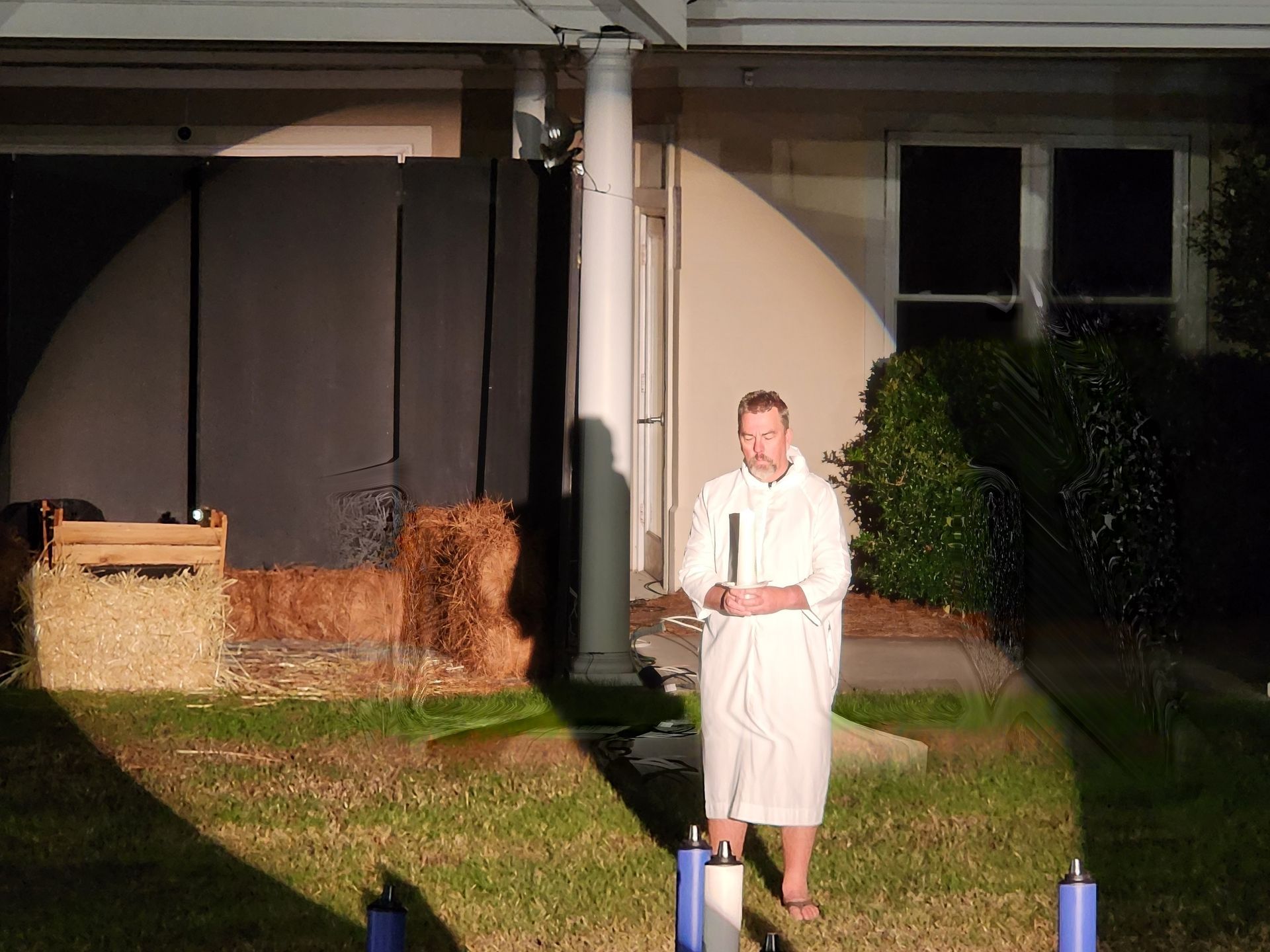 Man in white robe holds a lit candle. Stage set with hay bales, black backdrop, and a white column.
