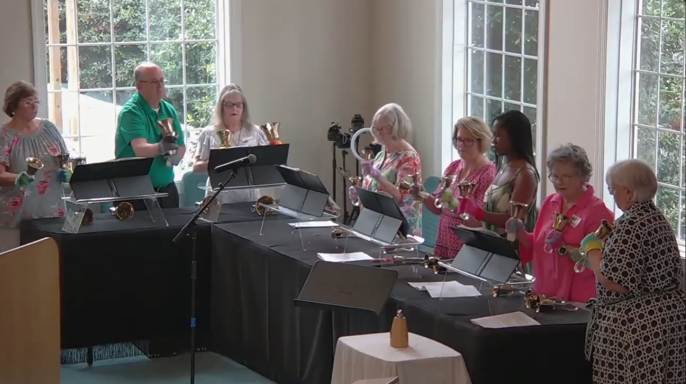 People in bright shirts perform with handbells in a sunlit room.