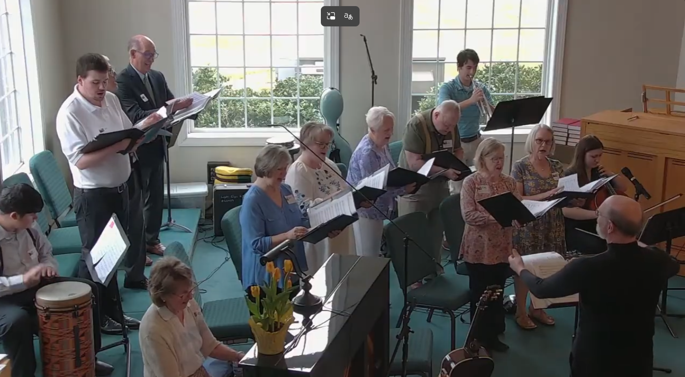 Choir group singing from sheet music in a bright room with large windows and a piano