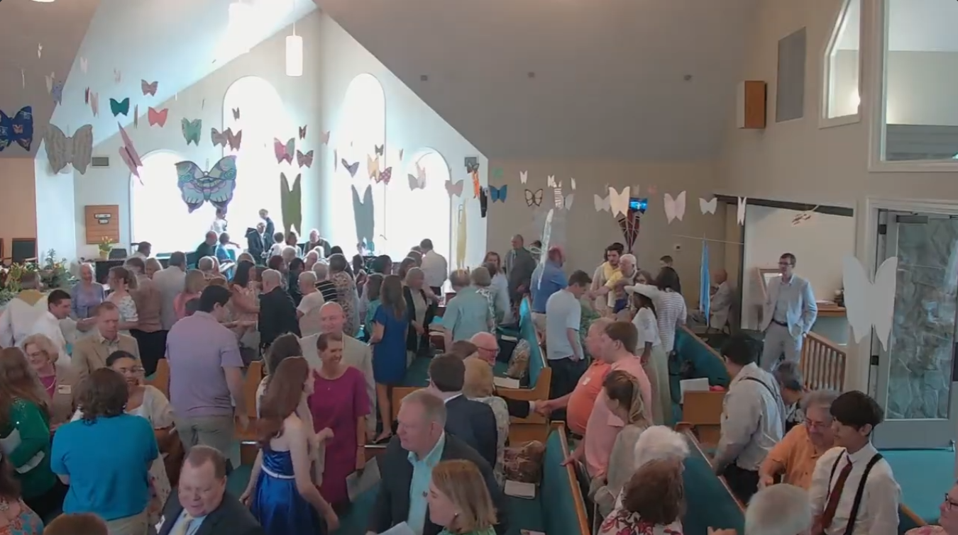Crowded indoor event with people seated and standing under hanging butterfly decorations and bright windows