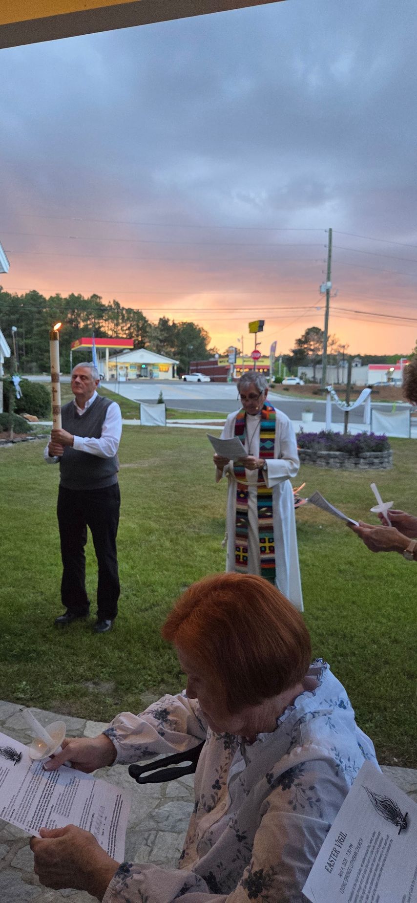 People in formal wear standing on a lawn at sunset, with chairs and a cloudy sky in the background