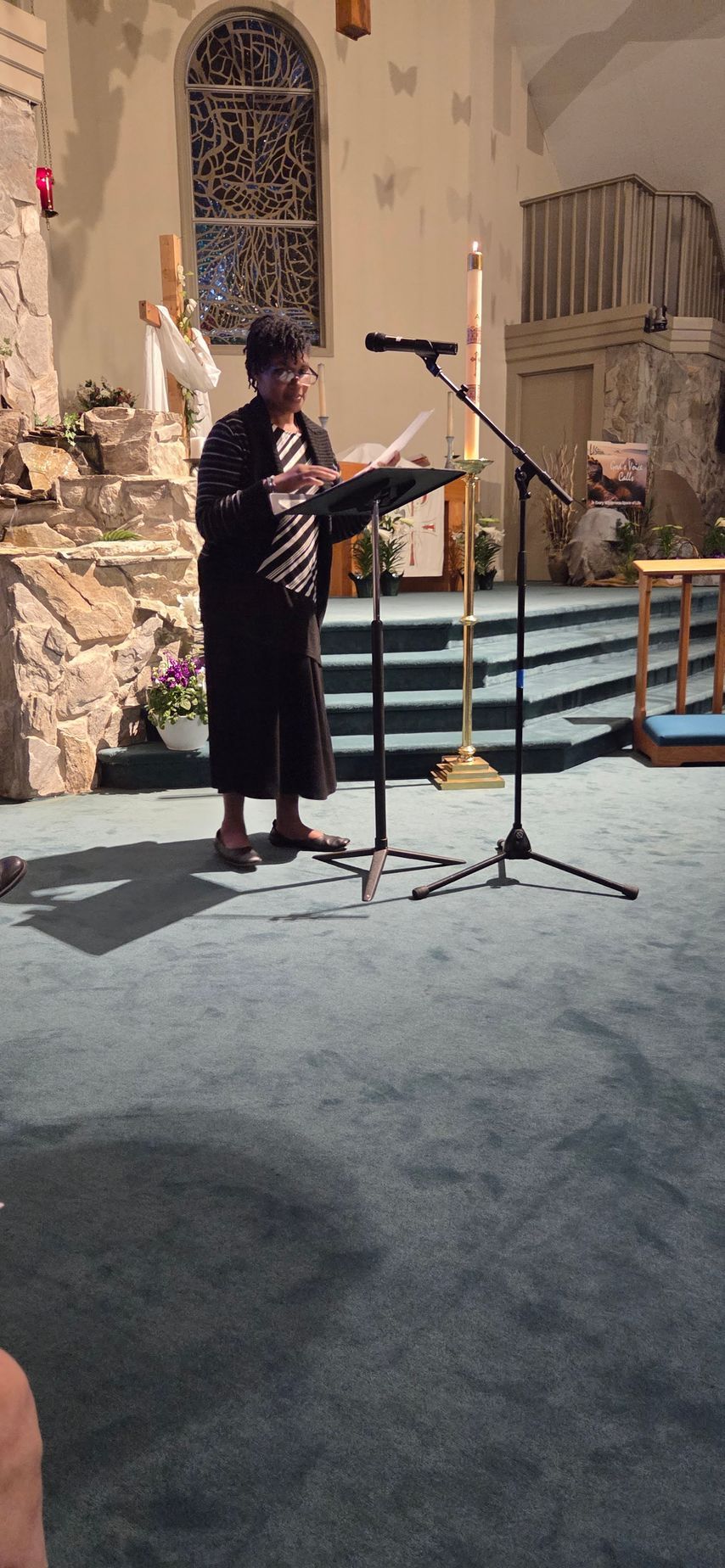 Person speaking at a lectern in a church, with microphone stands and altar steps in the background