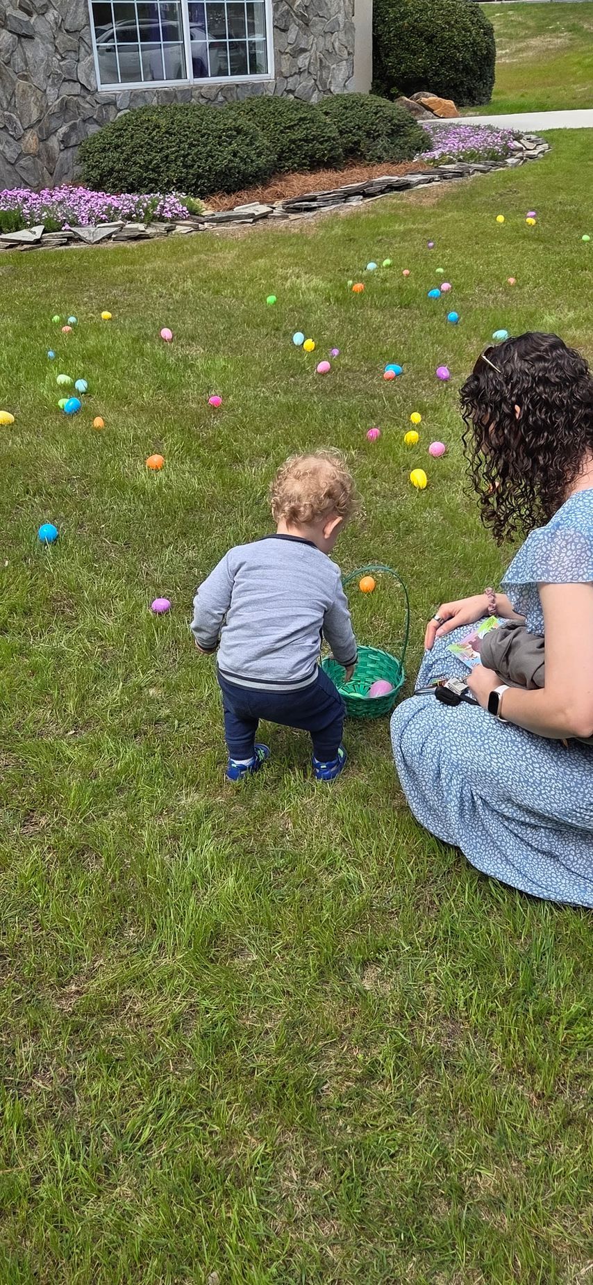 Adult and toddler sitting on grass near scattered colorful eggs in a yard