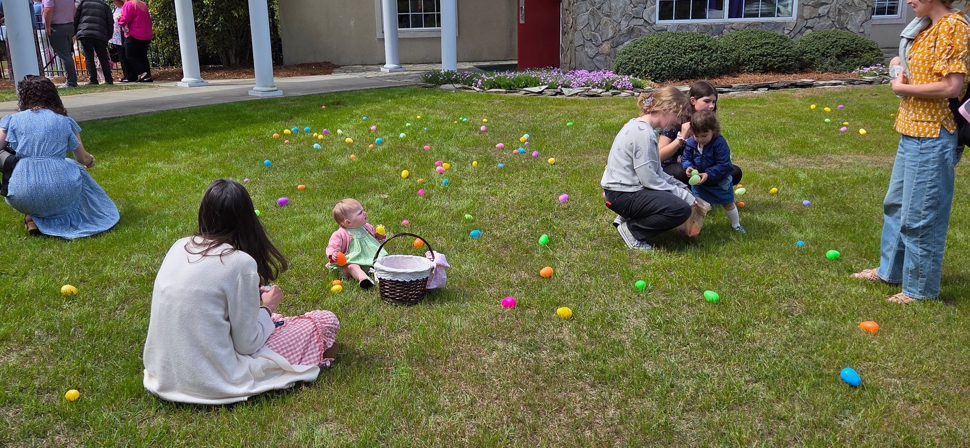 People on a grassy lawn with colorful plastic eggs, children playing outside near a building.