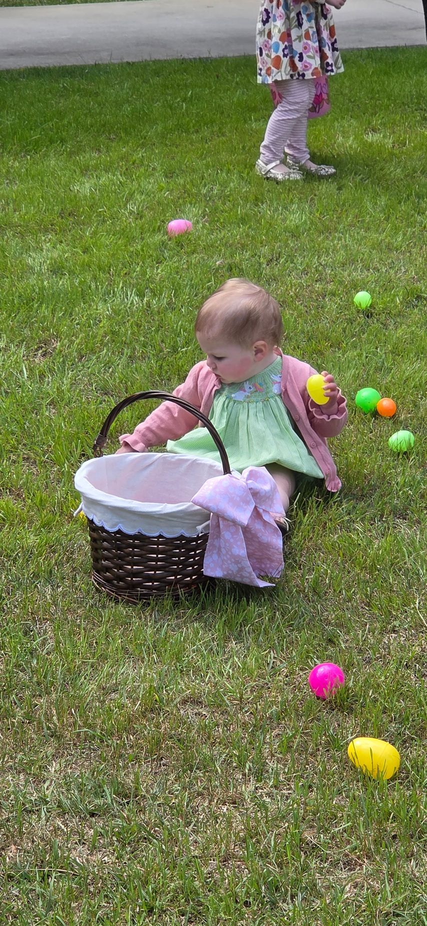 Toddler sitting on grass beside an Easter basket, reaching for colorful plastic eggs with more eggs scattered nearby