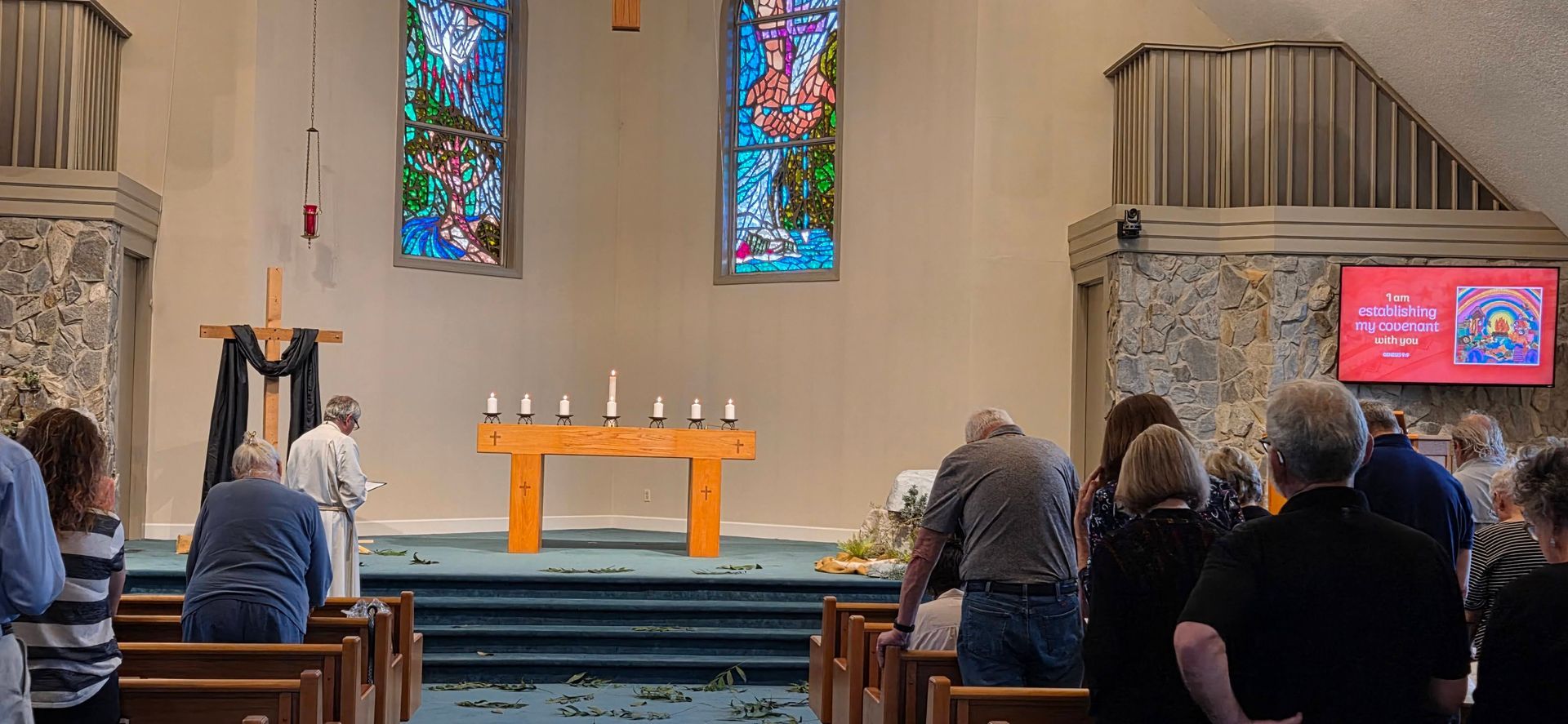 People seated in a church facing a lit altar with colorful stained-glass windows and candles