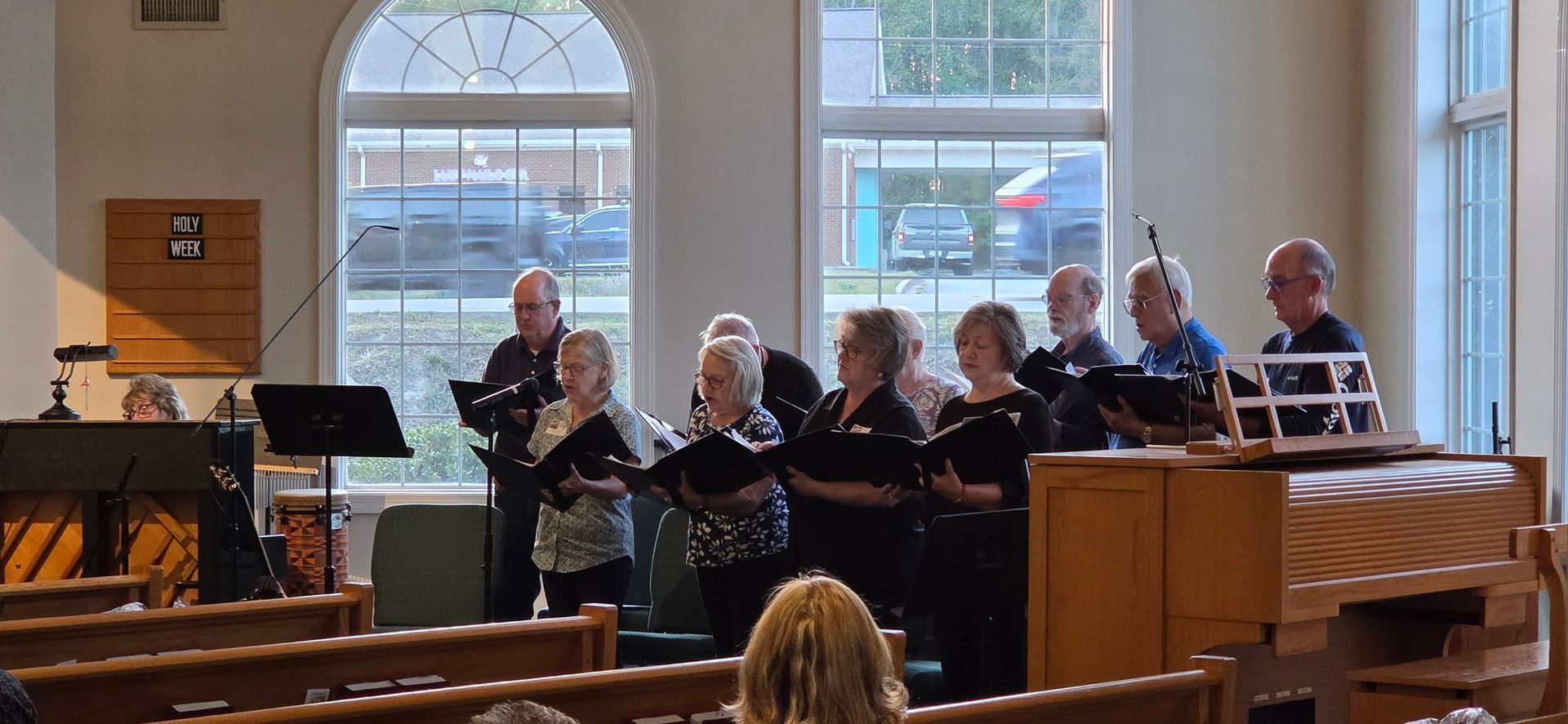 Choir singing in a church, standing before arched windows and a piano.