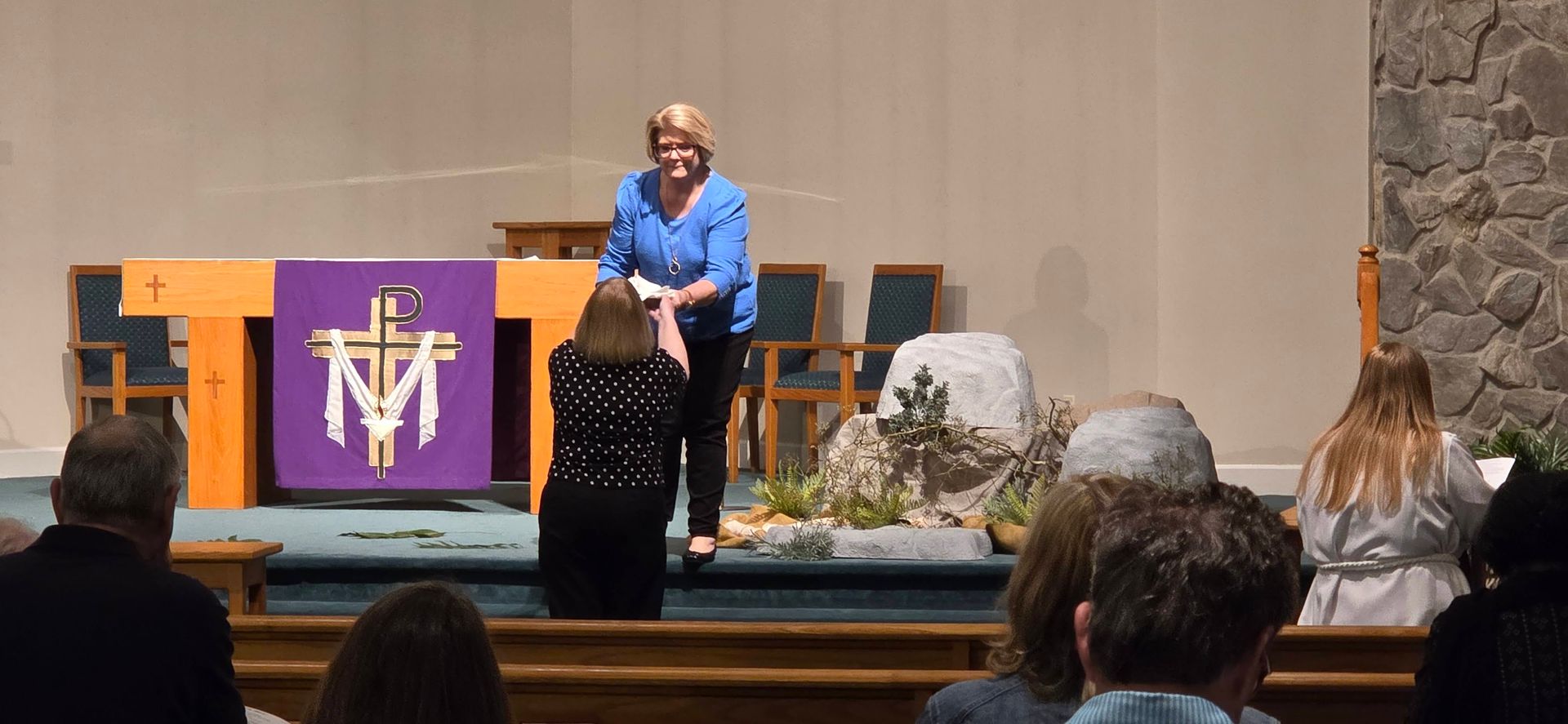 Woman in blue speaking at church podium with purple and orange cloths, audience seated in foreground